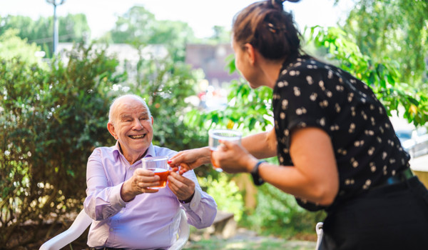 Older Man Sitting In Garden Smiling And Being Handed A Drink By A Woman