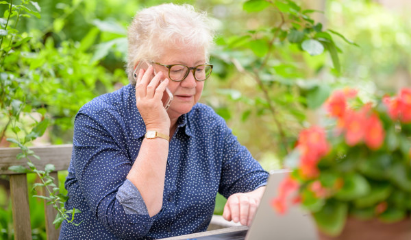 Older Woman In Garden Working On Her Laptop And On The Phone
