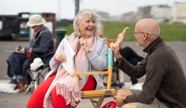 Older Woman Outside Sitting In Chair Smiling Chatting To Another Man