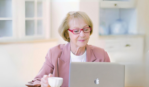 Older Woman With A Cup Of Tea Looking At Laptop On Kitchen Table